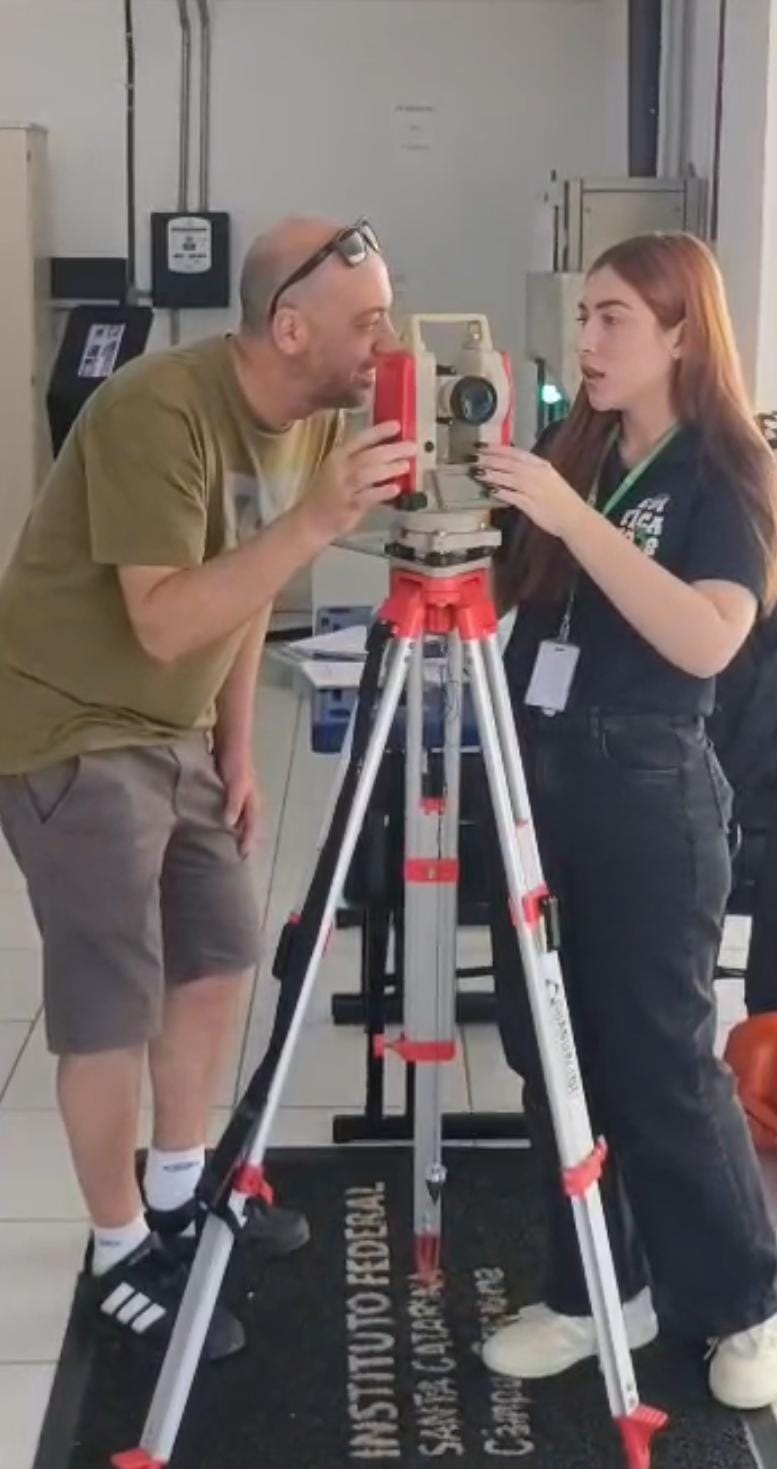 Aluna de Edificações apresentando para um visitante a funcionalidade do teodolito, um instrumento ótico de precisão usado na topografia e engenharia para medir ângulos horizontais e verticais