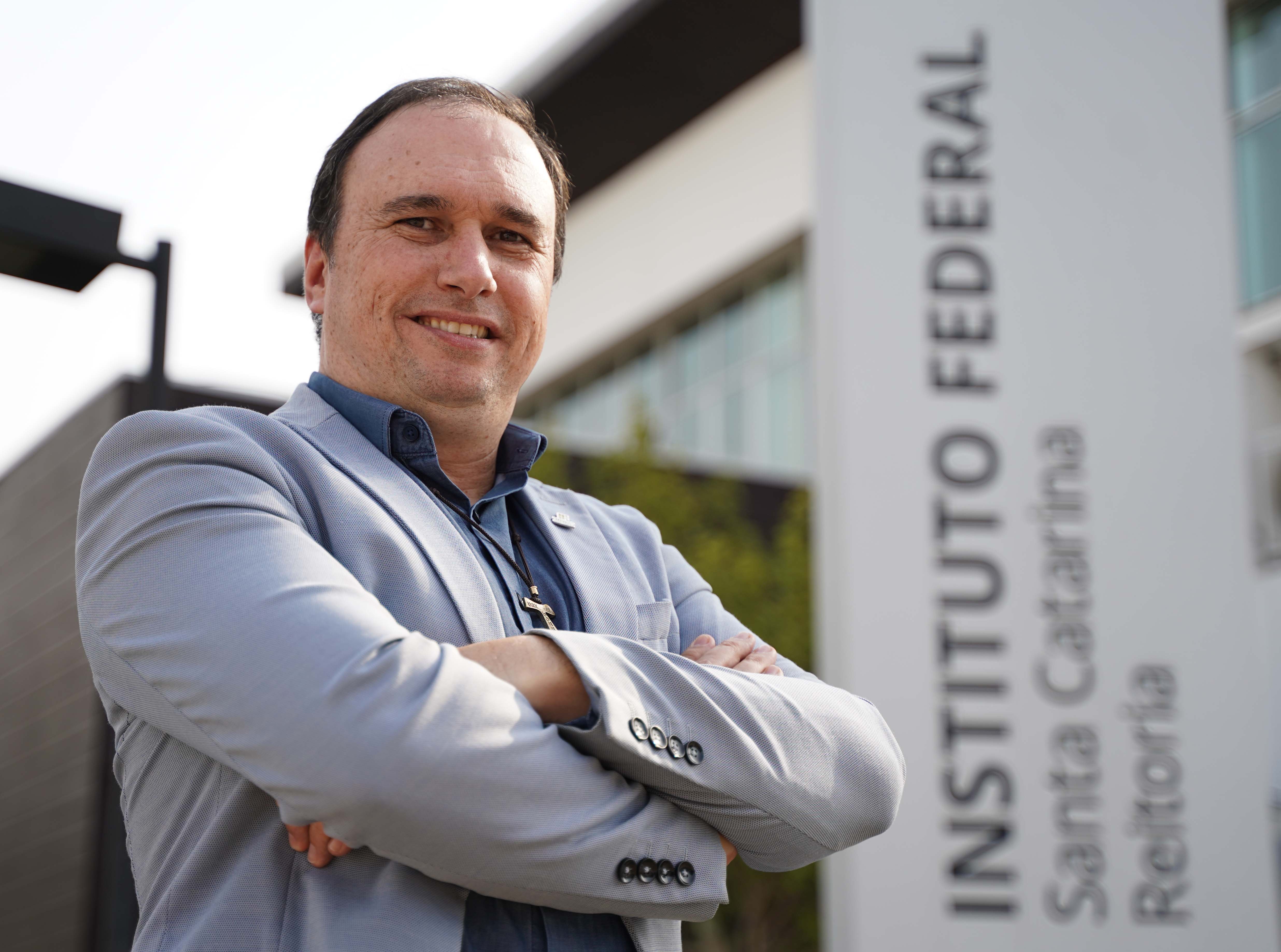 Um homem de pele clara, com cabelo escuro e curto, está sorrindo para a câmera. Ele usa um blazer cinza claro sobre uma camisa azul escura. Seus braços estão cruzados na frente do corpo. Ao fundo, parcialmente visível, há um prédio moderno com grandes janelas e uma placa vertical branca com o texto "INSTITUTO FEDERAL Santa Catarina Reitoria" em letras pretas. O céu está claro e o ambiente é externo.